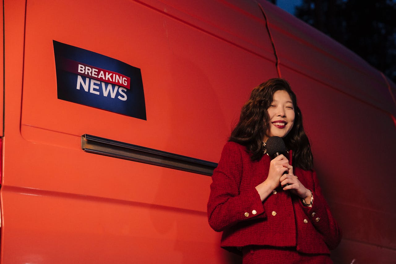 Asian female reporter smiling with microphone besides a news van at night.
